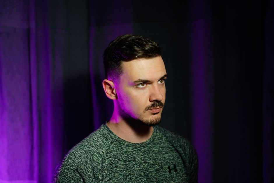 Studio portrait of a man against a dark backdrop with purple lighting, showcasing an artistic mood.