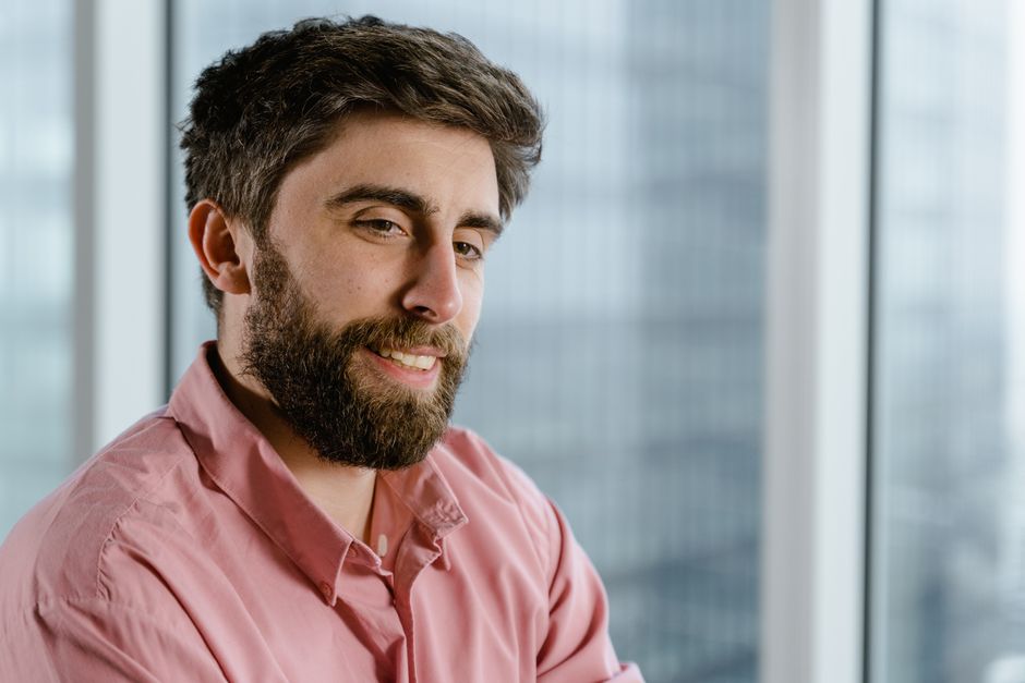 Portrait of a bearded man in a pink shirt smiling indoors with a blurred glass window background.